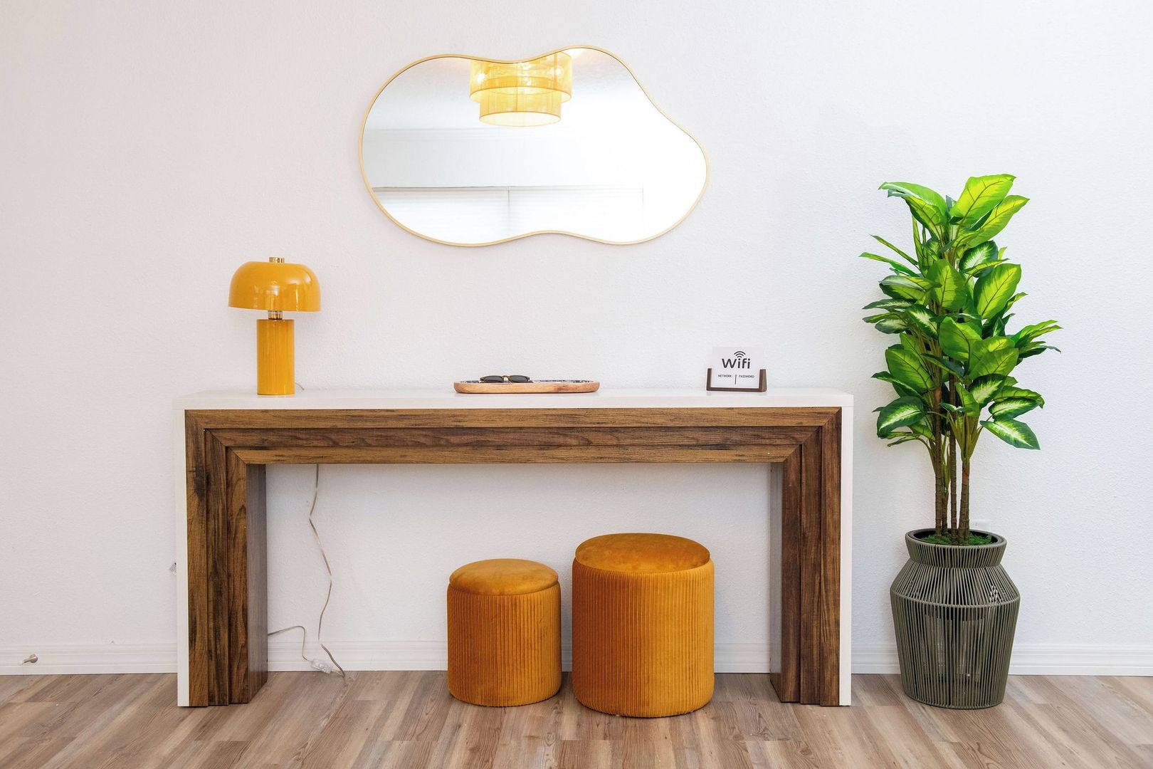A wooden console table with a mirror, two stools, a lamp, a potted plant, and a small sign.