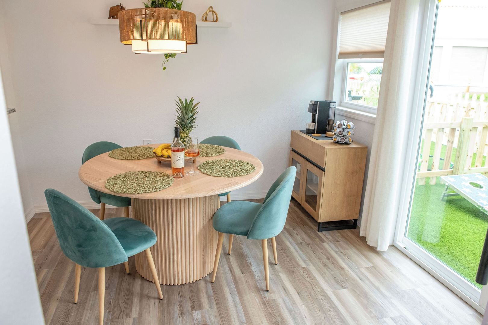 A dining area with a round wooden table, teal chairs, and a small kitchen area with a window.