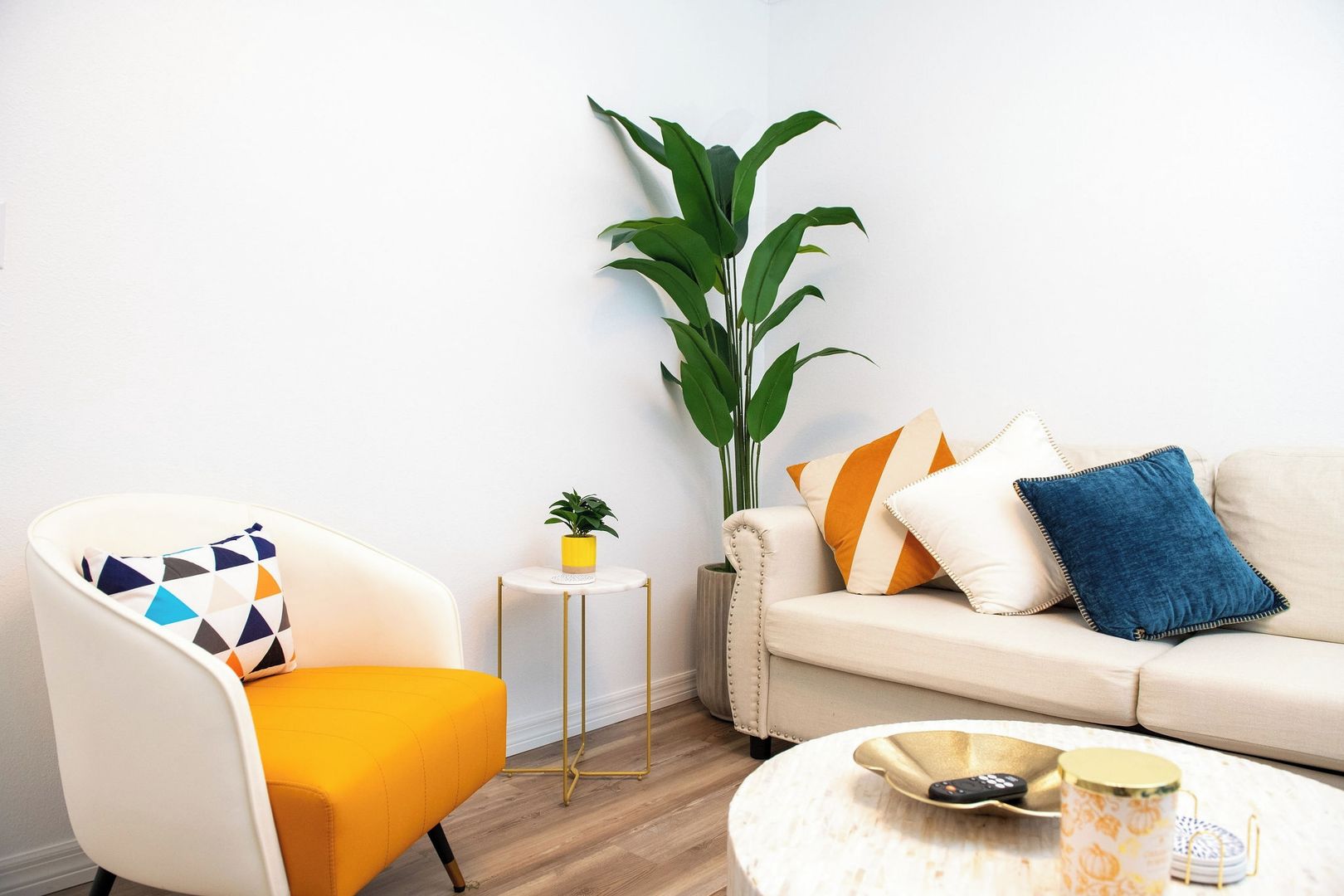 Een woonkamer met een gele fauteuil, een witte bank, een ronde salontafel en een potplant.