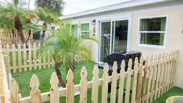 Een achtertuin met een houten omheining, een patio tafel, een barbecue en een kleine palmboom.