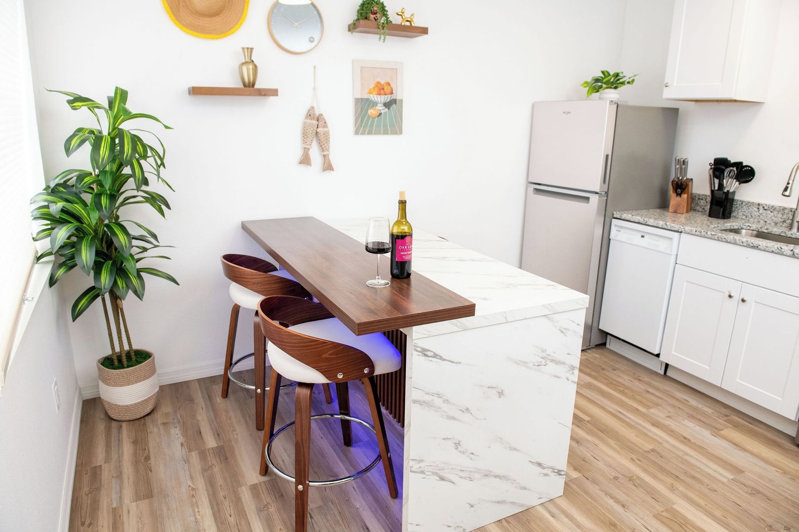 A modern kitchen with a marble island, wooden chairs, a wine bottle, and a potted plant.
