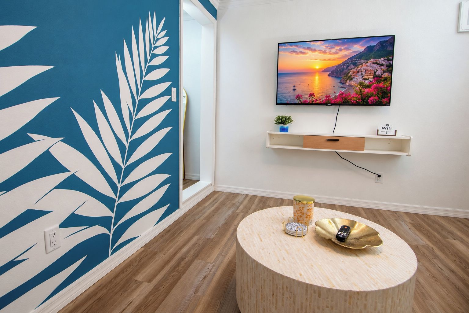 Una habitación con un gran mural en la pared, un televisor montado, una mesa redonda de madera y una estantería con una planta y un cartel.