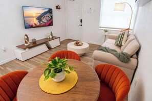 Ein modernes Wohnzimmer mit einem runden Holzesstisch, orangefarbenen Sesseln, einem beigen Sofa, einem Flachbildfernseher an der Wand und einem kleinen Beistelltisch mit einer Pflanze.