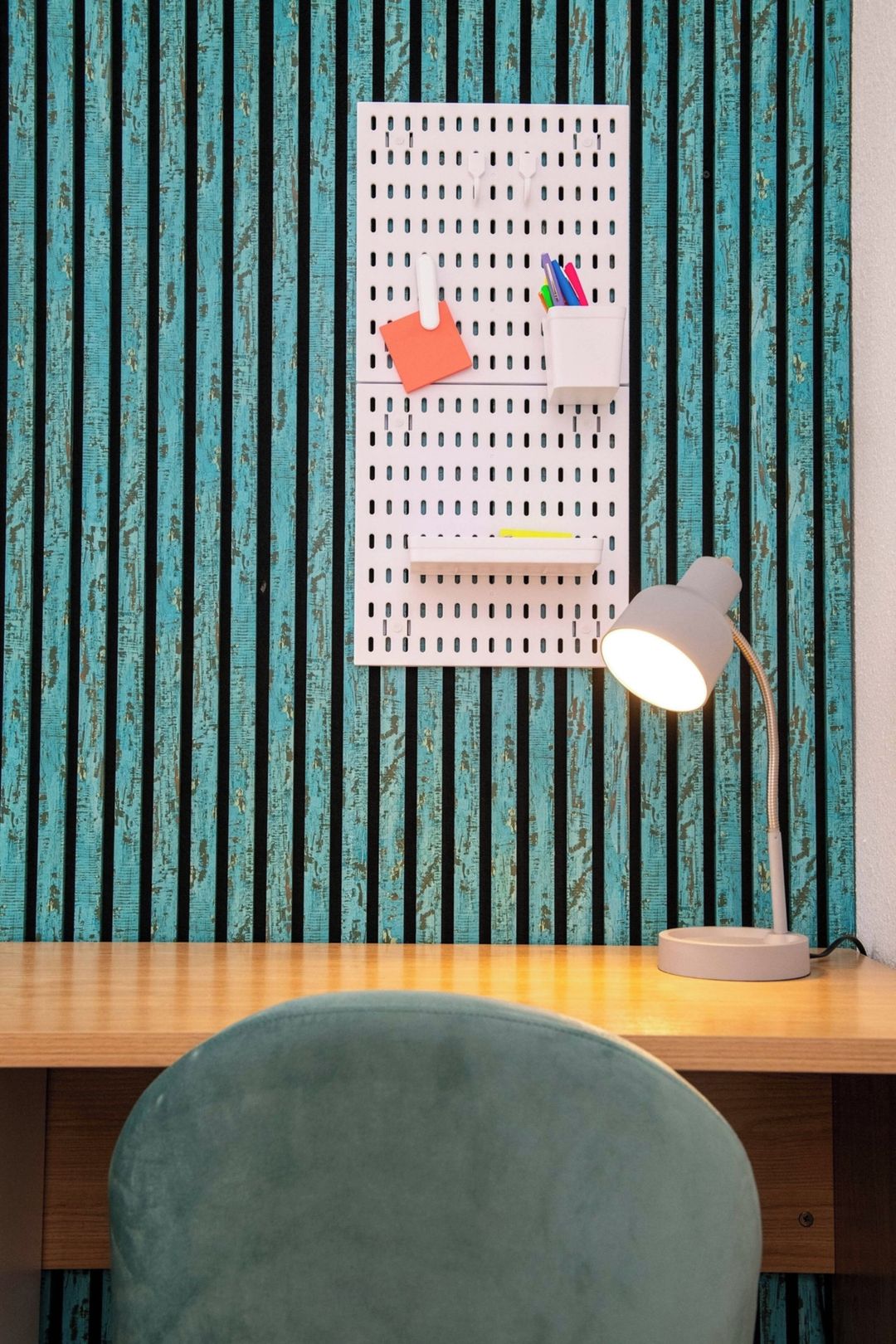 Un bureau avec un panneau perforé servant d’organisateur, une lampe et une chaise, le tout placé contre un mur à rayures.