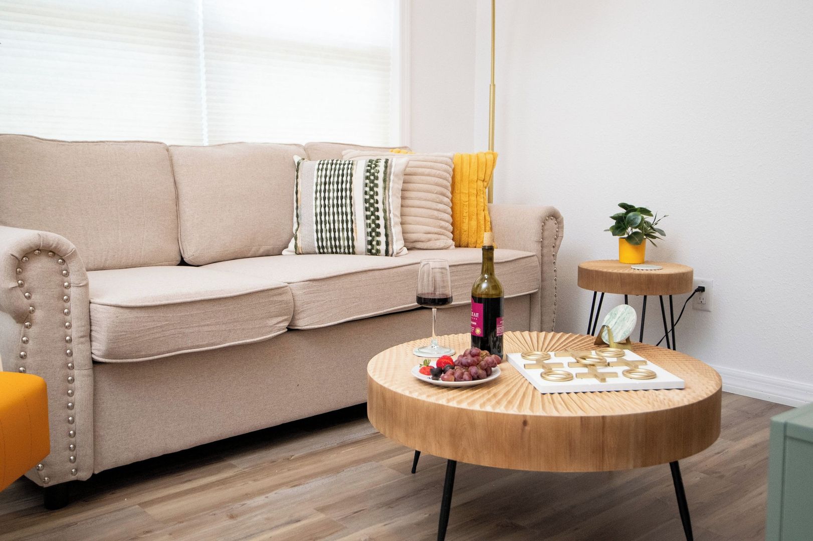 A living room with a beige sofa, a round wooden coffee table with snacks and a bottle, and a small side table with a plant.
