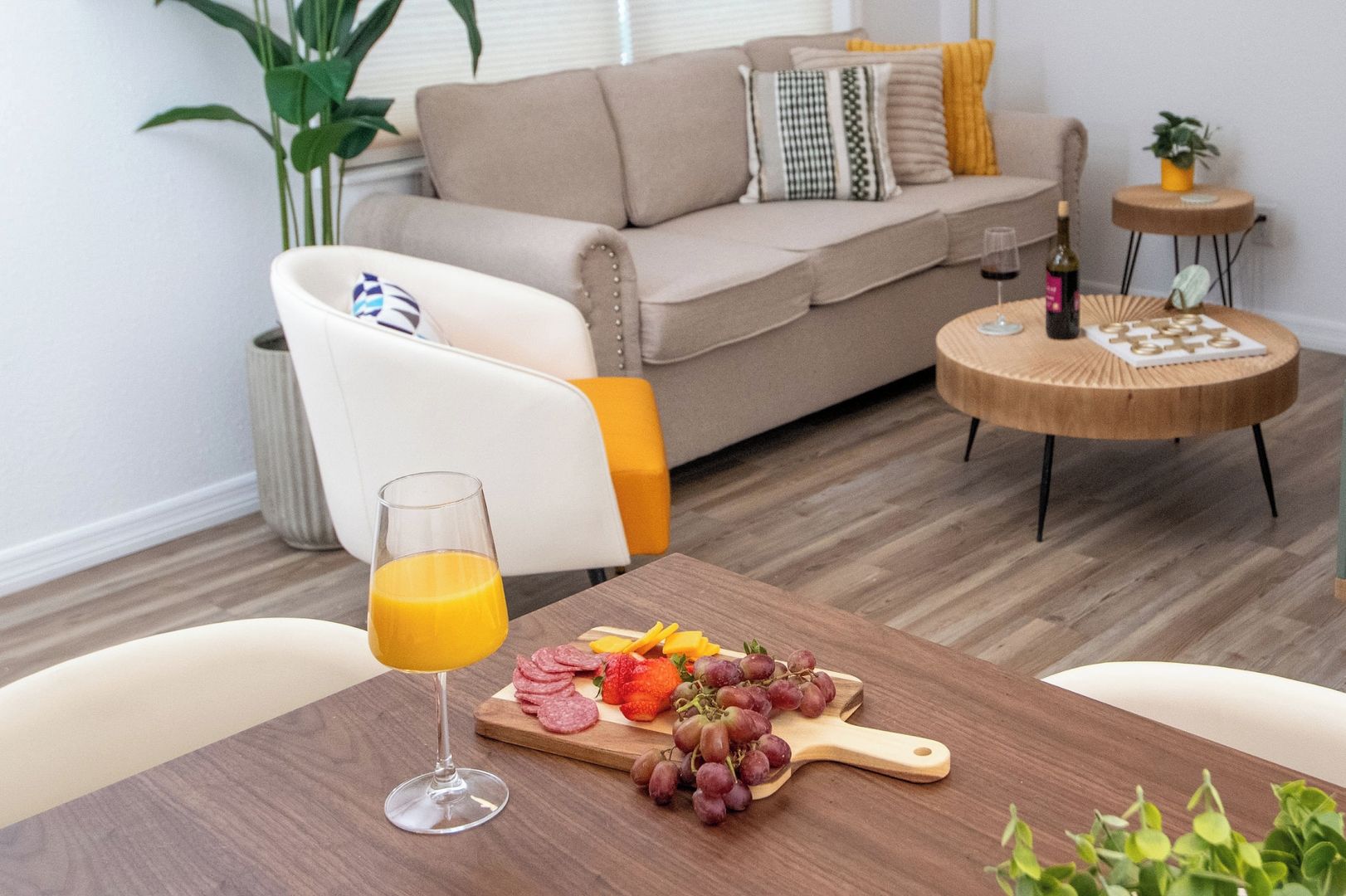 Una sala de estar moderna con una mesa de comedor de madera, un vaso de jugo de naranja, un plato con fiambres y uvas, un sofá y una pequeña mesita redonda con una botella.