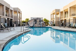 Ein Schwimmbecken mit einer Felsformation in der Mitte, umgeben von Liegestühlen und einem Gebäude mit Balkonen.