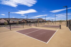 Una cancha de tenis exterior con una red, rodeada por una cerca, y casas al fondo.