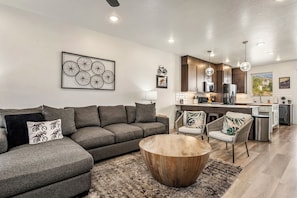 A modern living room with a sectional sofa, a round wooden coffee table, and a kitchen area with dark wood cabinets and stainless steel appliances. - Lofts 45, PS5, Balcony Views, and Resort Amenities! (Saint George)