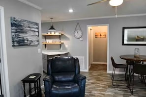 A modern living room with a black leather chair, a dining table with chairs, a kitchen area with a counter and shelves, and a clock on the wall.