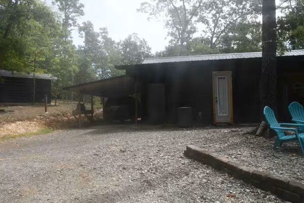Un chemin de gravier mène à un chalet rustique doté d’un toit en métal, d’une porte et d’une fenêtre. Deux chaises bleues sont placées à l’extérieur.