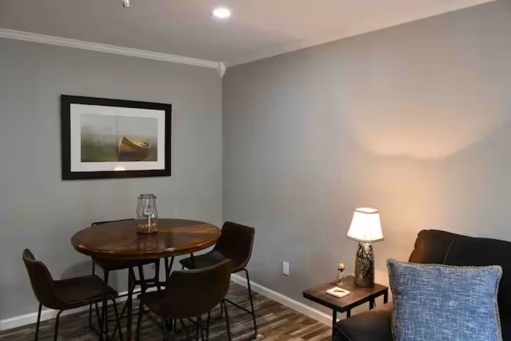 A dining area with a round table, four chairs, a framed picture on the wall, a small table with a lamp, and a blue pillow on a couch.