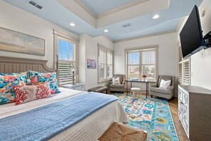 A bedroom with a bed, two chairs, a small table, a TV mounted on the wall, and large windows with blinds. - Sea Gate #102, This 1 bedroom suite on St. Simons (St. Simons Island)