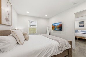 A bedroom with a bed, pillows, a window with blinds, a painting on the wall, and a door leading to another room. - Two Private Homes | 5 King Bedrooms | Sleeps 10 (Lafayette)