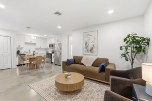 A modern living room with a brown sofa, a round coffee table, and a dining area with wicker chairs. - Two Private Homes | 5 King Bedrooms | Sleeps 10 (Lafayette)