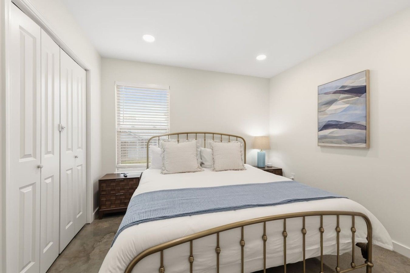 A bedroom with a bed, a nightstand, a window with blinds, a painting on the wall, and a closet.