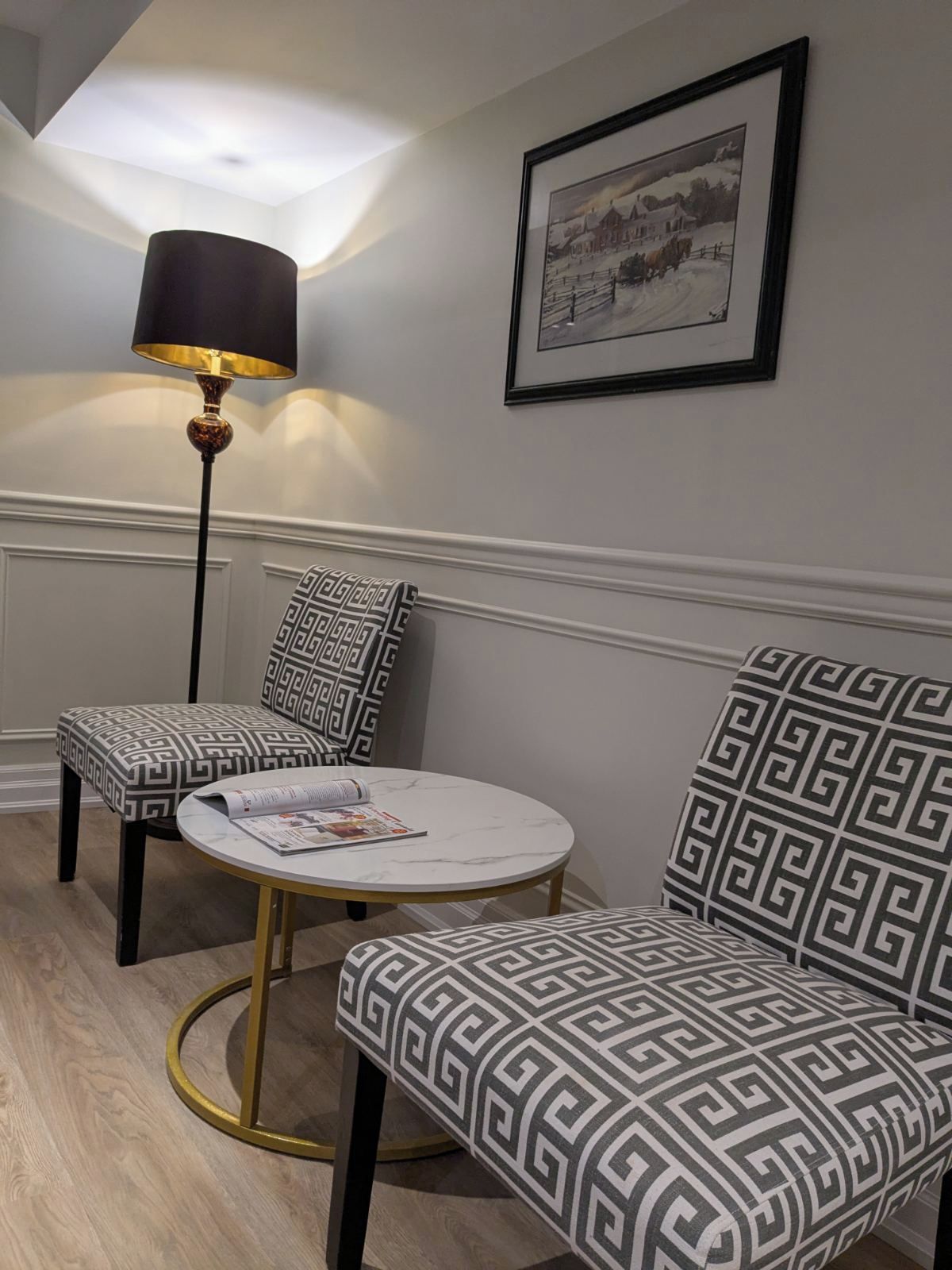 A room with a round table, two patterned chairs, a floor lamp, and a framed picture on the wall.