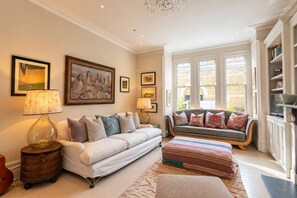 Ein helles Wohnzimmer mit einem weißen Sofa, einem gestreiften Hocker und einer gepolsterten Bank. An den Wänden befinden sich mehrere gerahmte Bilder, auf einem Holztisch steht eine Lampe und eine Bücherwand ist mit Büchern gefüllt.