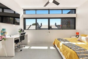 Ein modernes Schlafzimmer mit Bett, Schreibtisch und Stuhl, großen Fenstern und einem Deckenventilator.