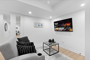 Un salon moderne avec un canapé gris, des coussins rayés noirs et blancs, une petite table basse et un téléviseur fixé au mur diffusant un film.