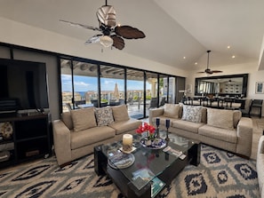 A spacious living area with a large flat-screen TV, a sectional sofa, a glass coffee table with decorative items, and a ceiling fan. - Beautiful "Villa De Familia" Partial Ocean View (Baja California Sur)
