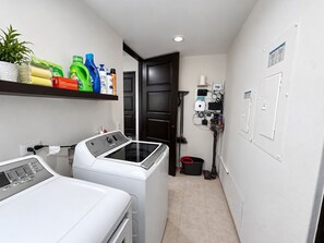 A laundry room with two front-loading washing machines, a shelf with cleaning supplies, and a wall-mounted electrical panel. - Beautiful "Villa De Familia" Partial Ocean View (Baja California Sur)