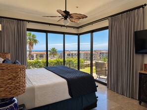 A bedroom with a large bed, a ceiling fan, a TV, and a view of a residential area. - Beautiful "Villa De Familia" Partial Ocean View (Baja California Sur)