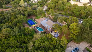 An aerial view of a residential property with a swimming pool, tennis court, and a large house surrounded by dense trees.