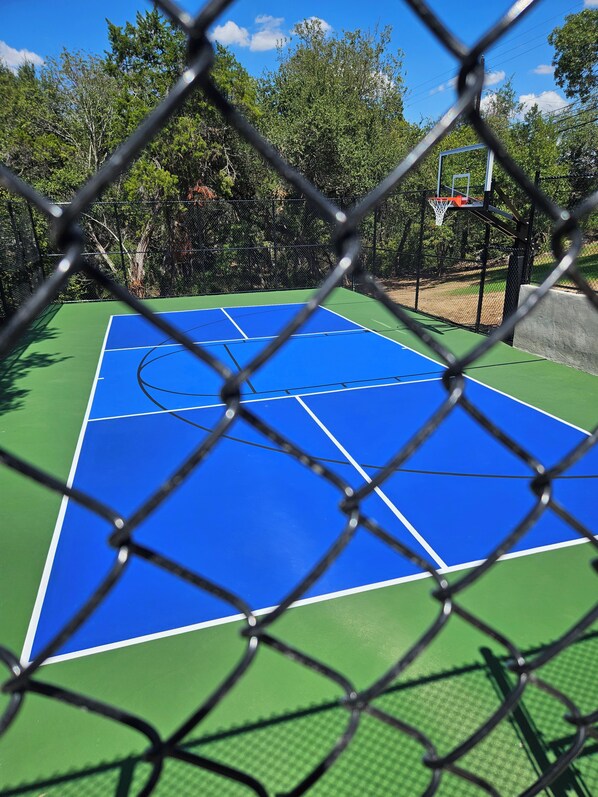 A chain-link fence surrounding a blue tennis court with a basketball hoop. - Toro Haus: Secluded Pool & Pickleball Paradise (Austin)