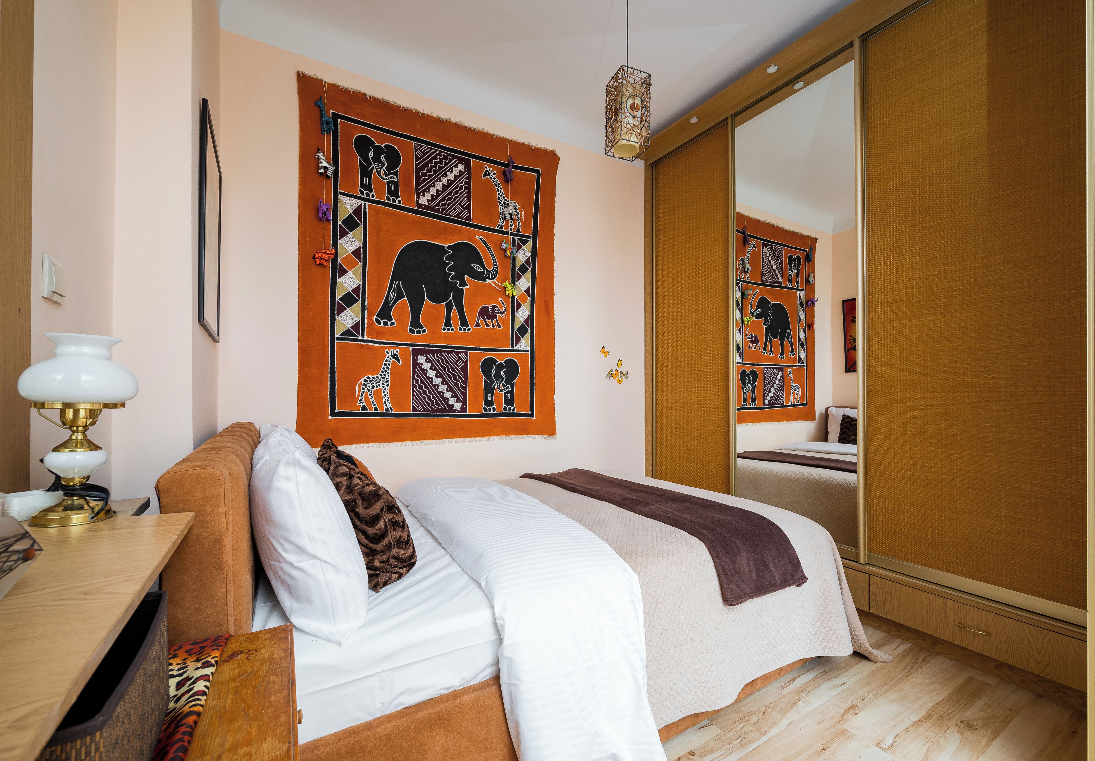 A hotel room with a bed, a bedside table with a lamp, a large mirror, and a wall tapestry depicting animals.