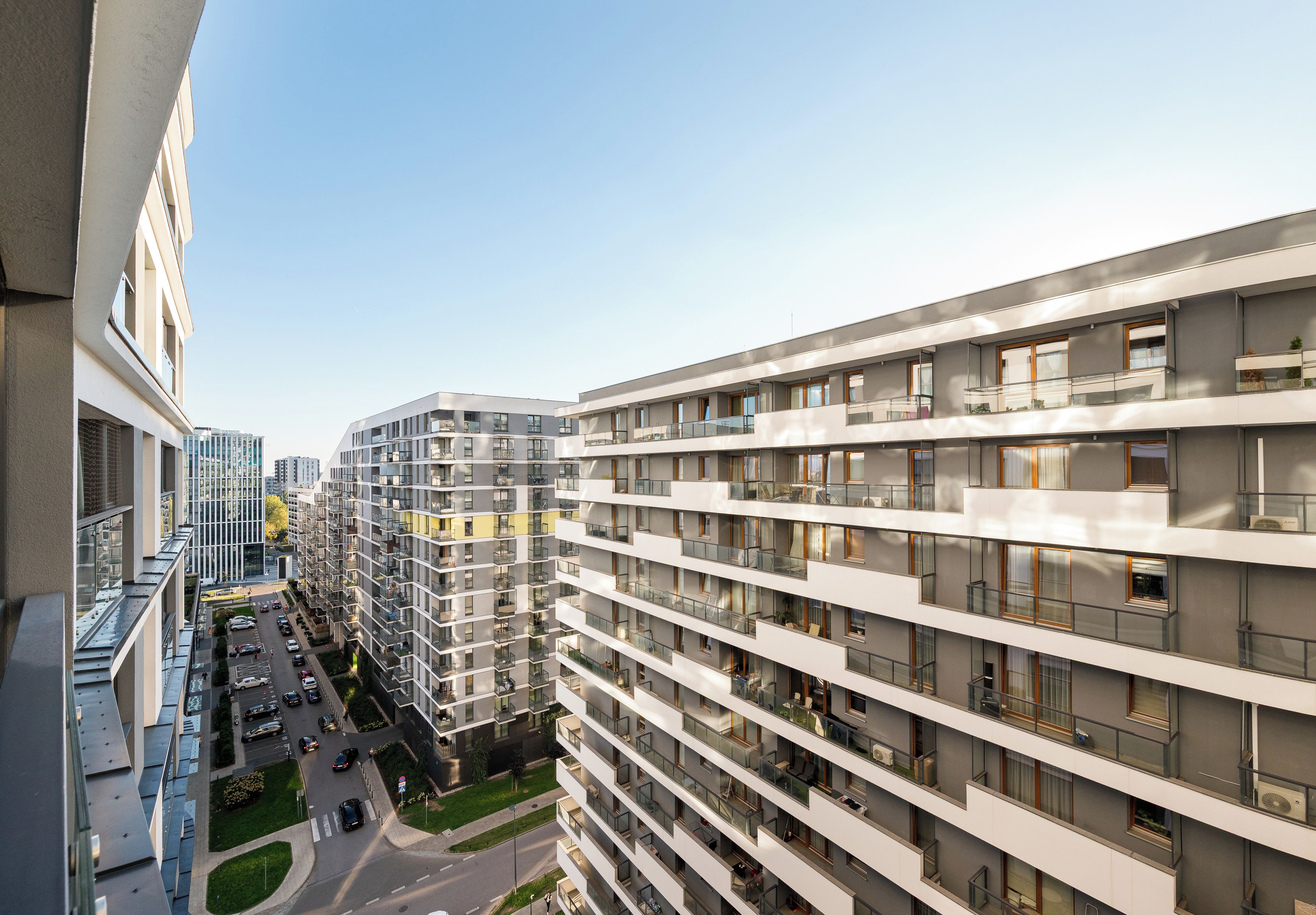 A multi-story residential building with balconies, a parking area, and a clear sky.