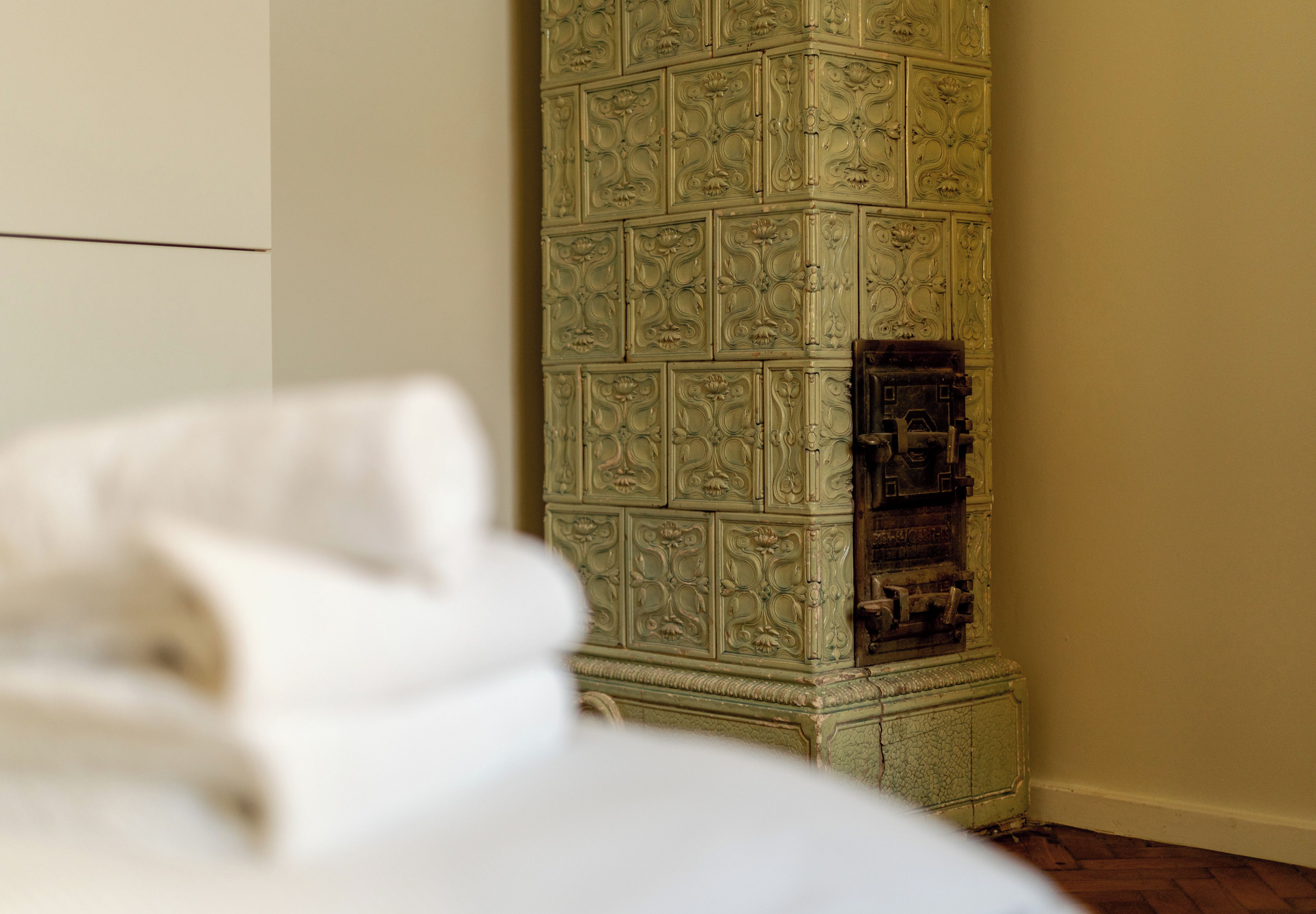 Un angolo di una stanza con un armadietto verde e decorato appoggiato al muro, un letto bianco con coperta bianca e un pavimento in legno.