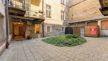 A courtyard with a paved floor, a small garden area with green plants, and a building with a wooden door and windows.