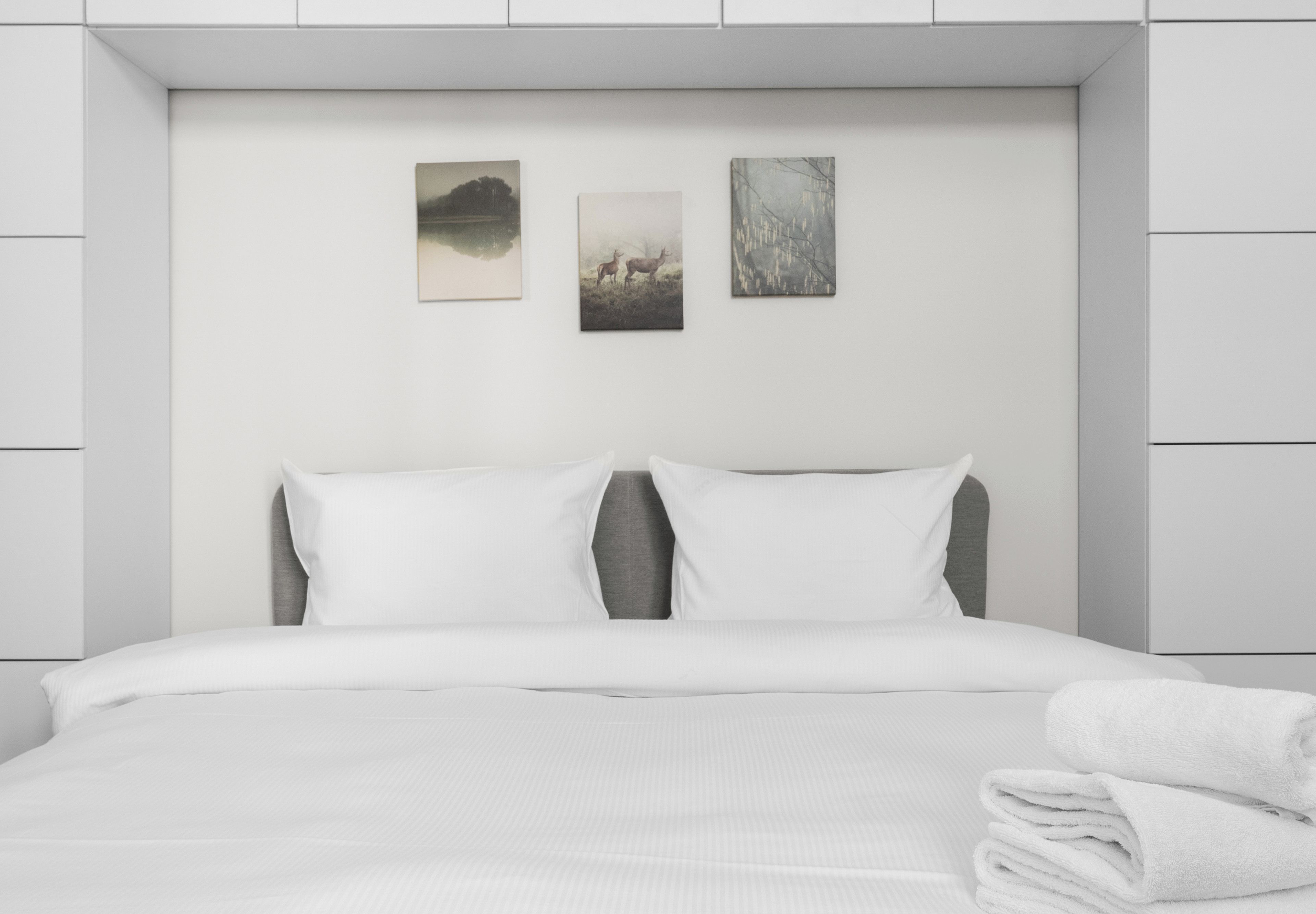 A modern hotel room with a neatly made bed, two pillows, and a stack of white towels. There are three framed artworks on the wall above the bed.