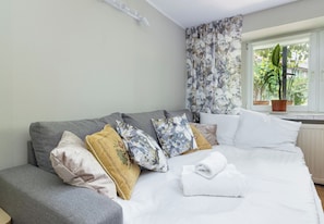 Een netjes opgemaakt bed met witte lakens, een grijze bank met veel kussens en een raam met uitzicht op groen.