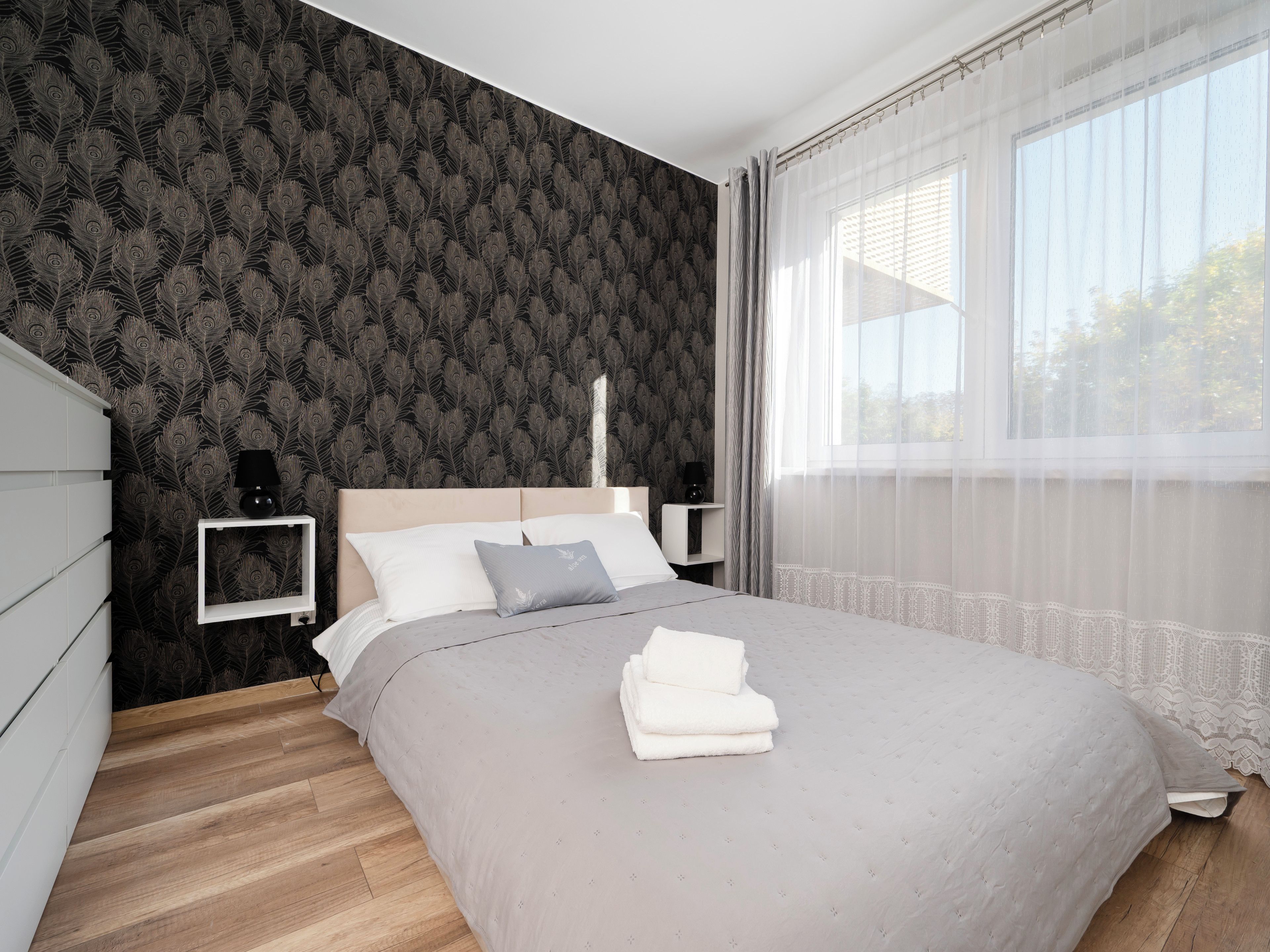 Een net opgemaakt bed met kussens en een opgevouwen handdoek, een nachtkastje met een lamp, en een raam met dunne gordijnen.