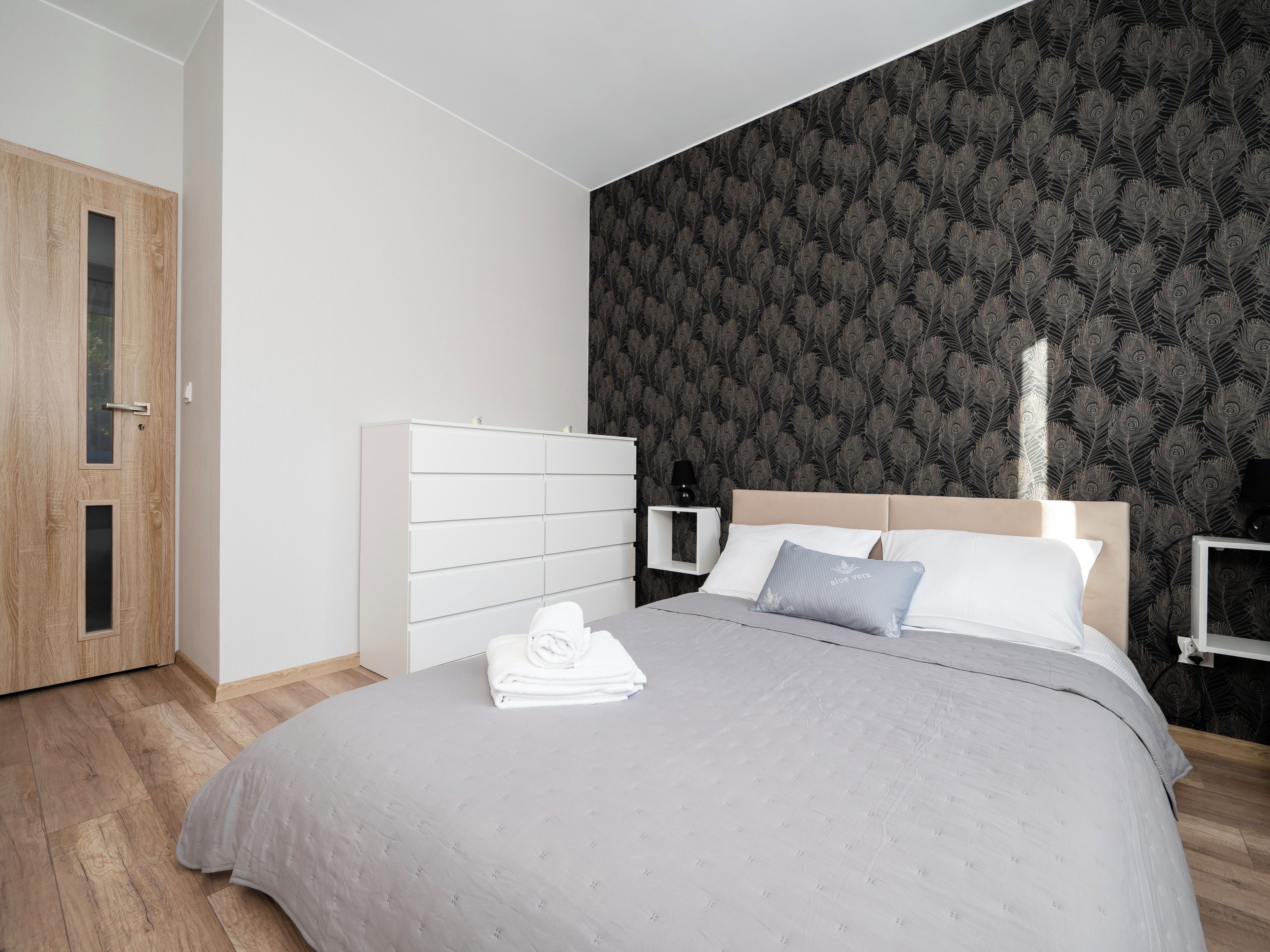 A bedroom with a bed, a white dresser, a nightstand, and a dark patterned wall.