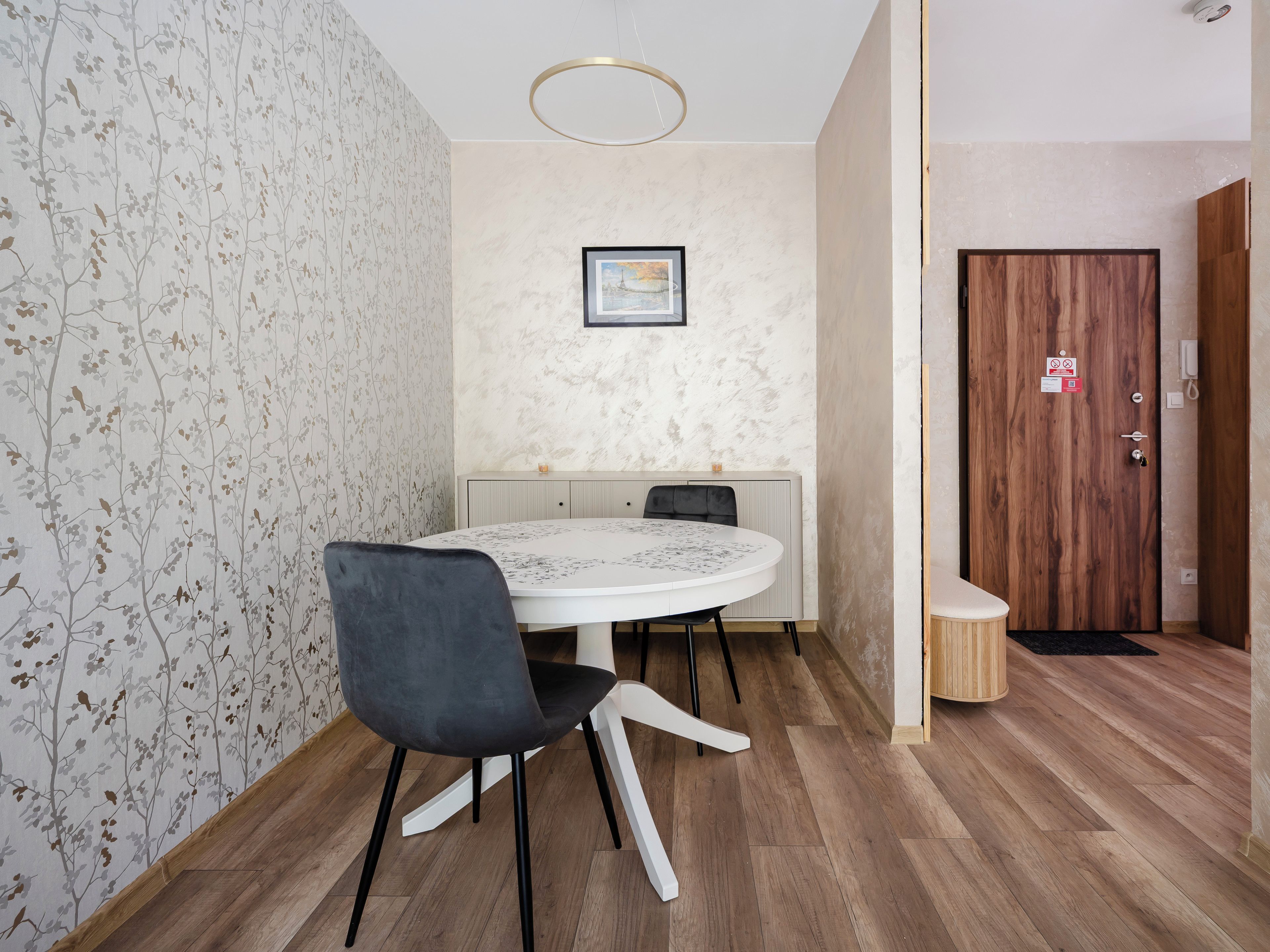 A room with a round dining table, a single chair, a framed picture on the wall, a wooden door, and a patterned wallpaper.