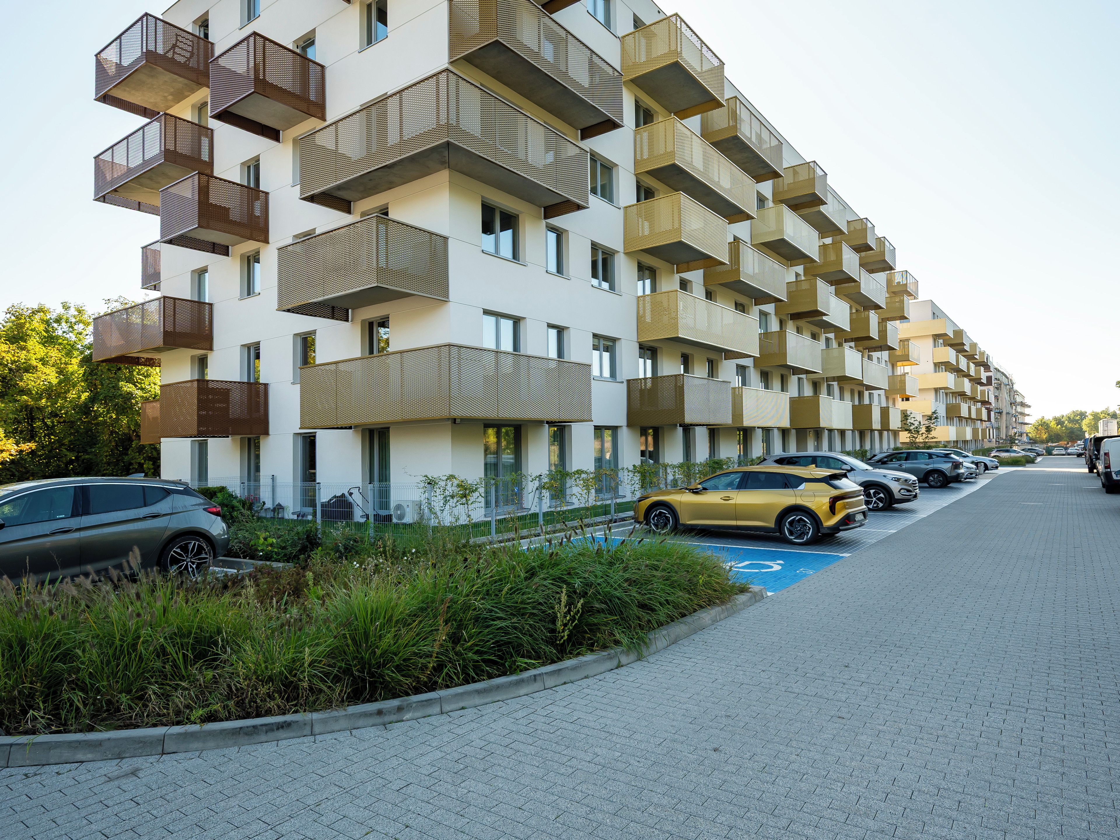 A multi-story residential building with balconies, a parking area, and a clear sky.