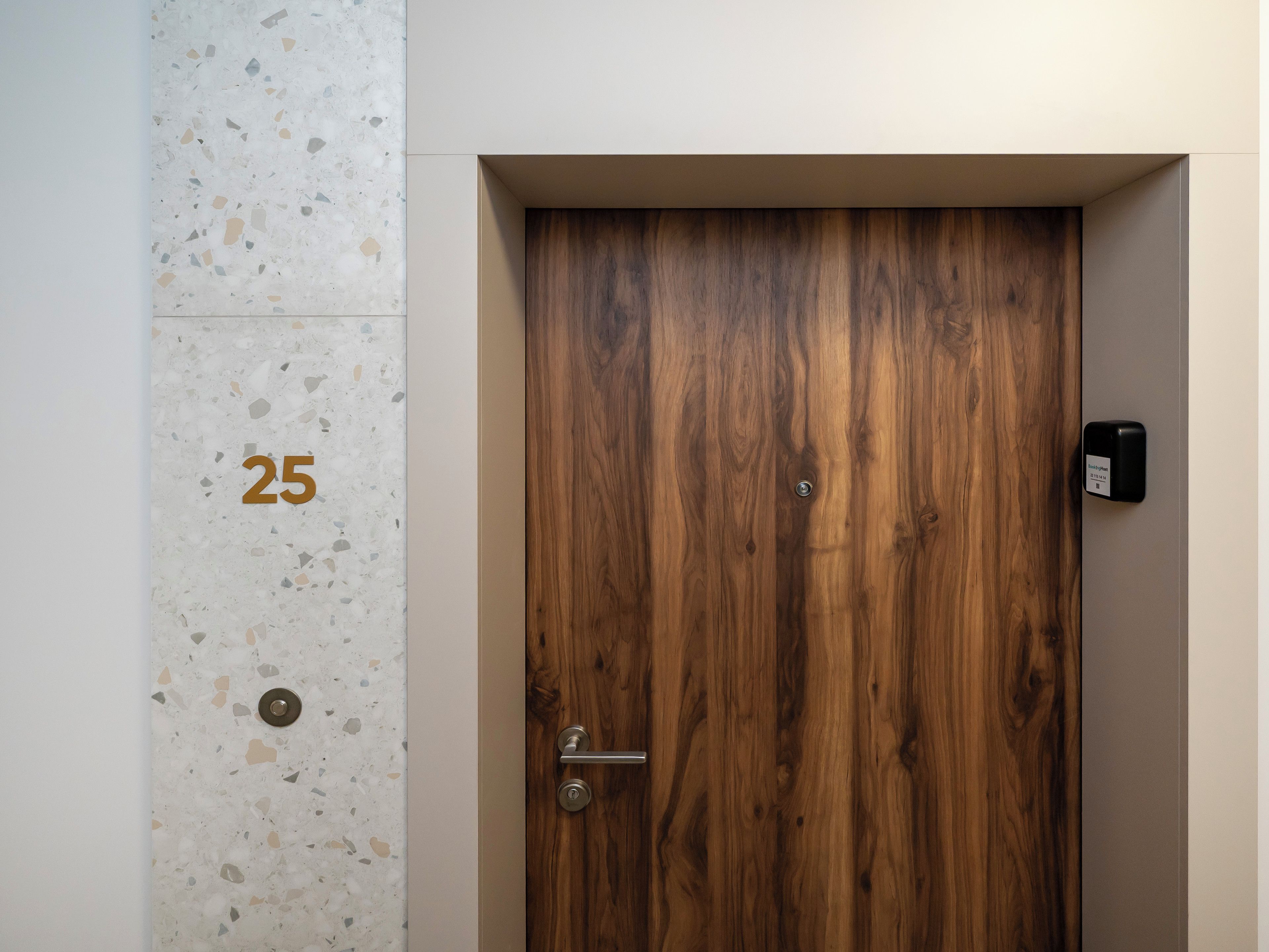 Una puerta de madera con un picaporte metálico y una cerradura, el número '25' en la pared, y un objeto negro pequeño a la derecha.