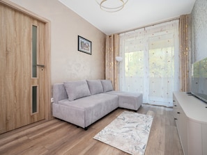 Ein modernes Wohnzimmer mit grauem Sofa, Holzboden, einem gerahmten Bild an der Wand und einem großen Fenster mit Vorhängen.
