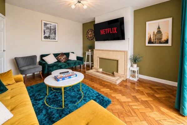 A living room with a green sofa, a grey armchair, a coffee table, a fireplace, and a flat-screen TV mounted on the wall. - Spacious 4BR Sleeps 10 Free Parking (Cardiff)