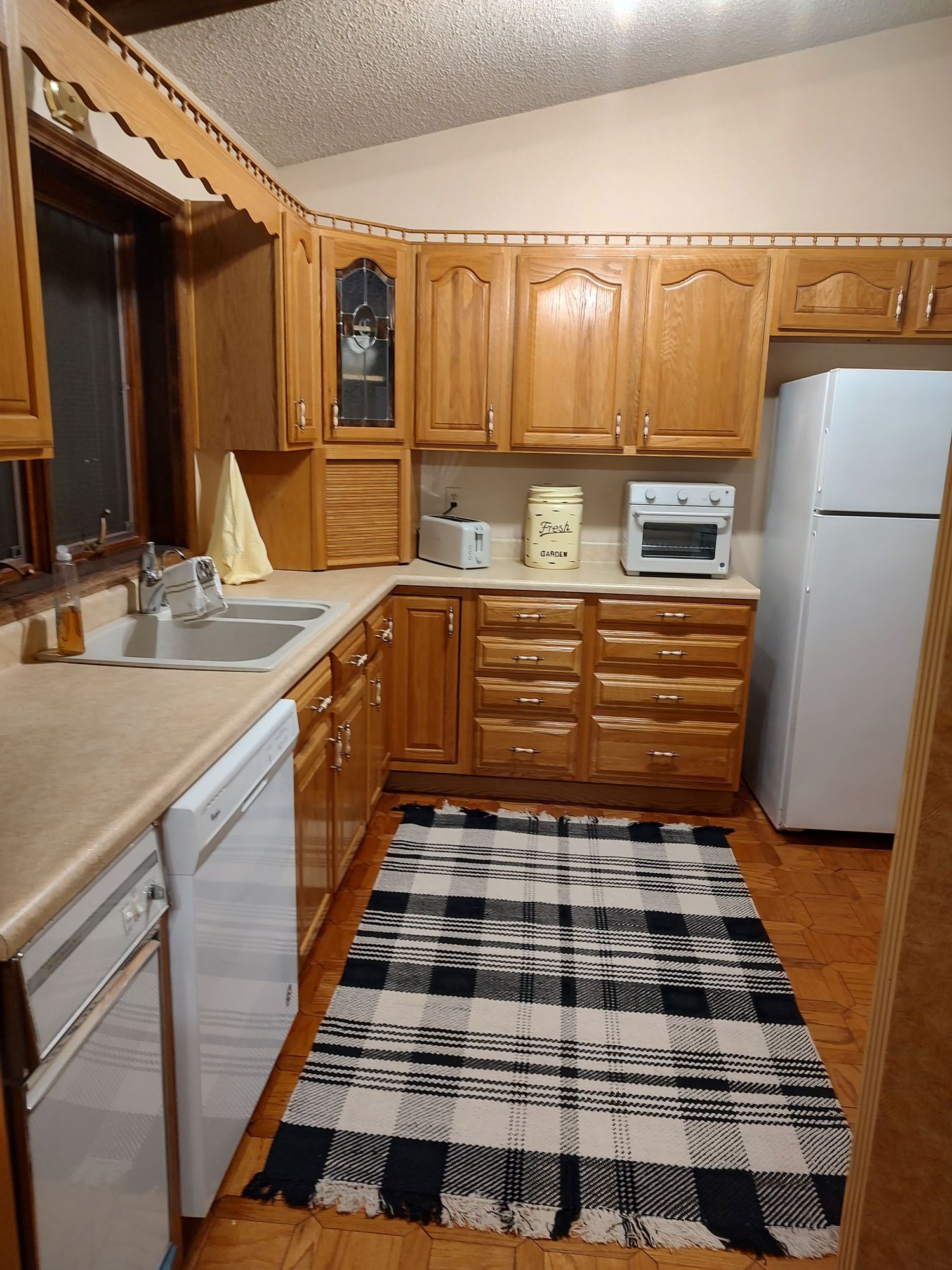 Une cuisine compacte avec des meubles en bois, un évier, un grille-pain, un réfrigérateur et un tapis à carreaux.