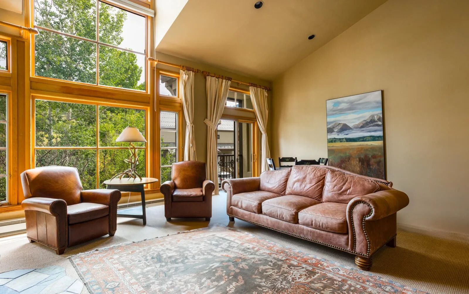 A spacious room with large windows, a brown leather sofa, two armchairs, a small table with a lamp, and a large painting on the wall.