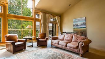 A spacious room with large windows, a brown leather sofa, two armchairs, a small table with a lamp, and a large painting on the wall.