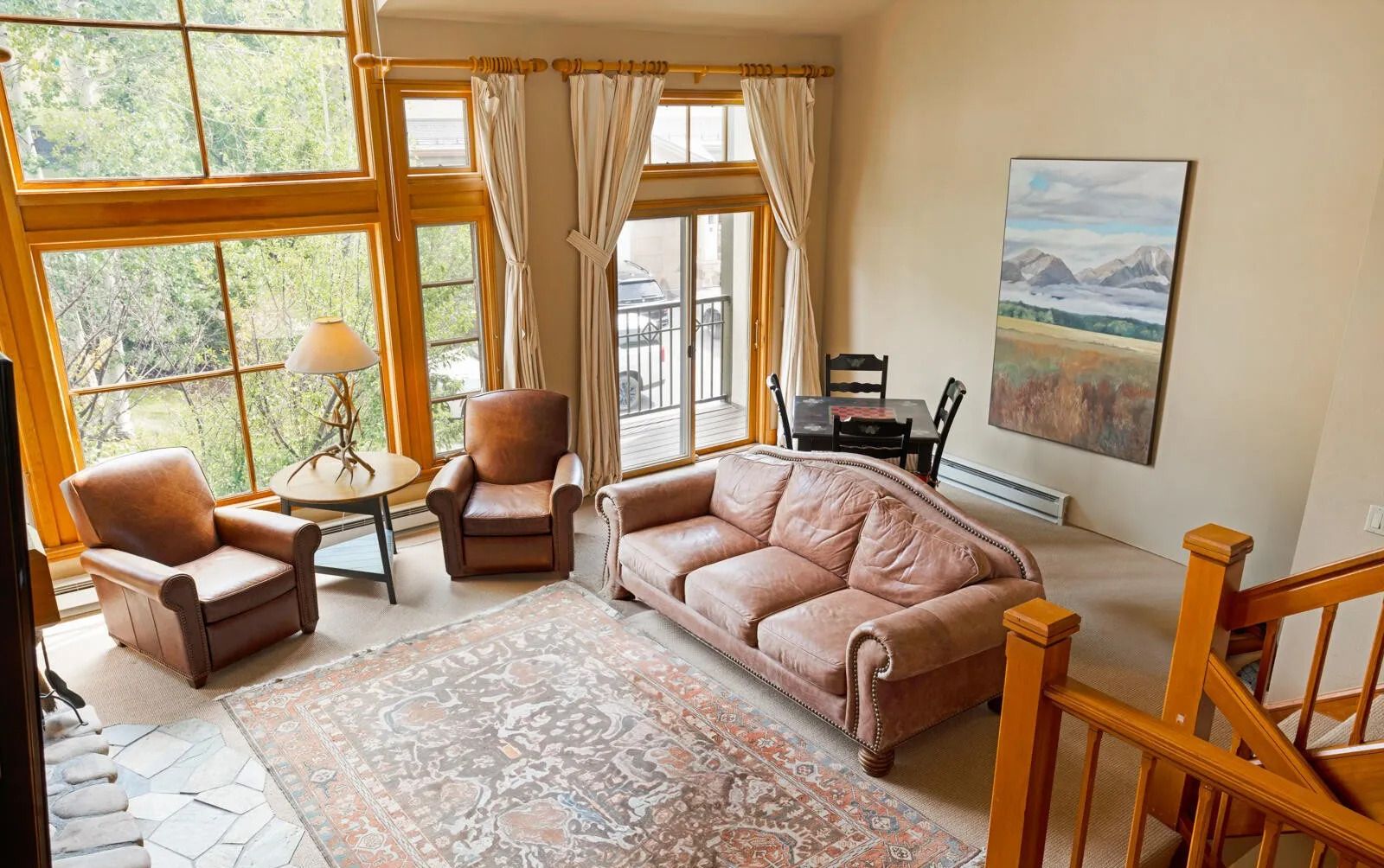 Un salon chaleureux avec un canapé en cuir, des fauteuils, une table basse et un tapis à motifs. De grandes fenêtres, ornées de rideaux, laissent entrer la lumière naturelle et offrent une vue sur la verdure environnante.