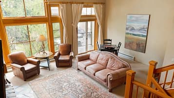 Un salon chaleureux avec un canapé en cuir, des fauteuils, une table basse et un tapis à motifs. De grandes fenêtres, ornées de rideaux, laissent entrer la lumière naturelle et offrent une vue sur la verdure environnante.