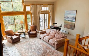 Un salon chaleureux avec un canapé en cuir, des fauteuils, une table basse et un tapis à motifs. De grandes fenêtres, ornées de rideaux, laissent entrer la lumière naturelle et offrent une vue sur la verdure environnante.
