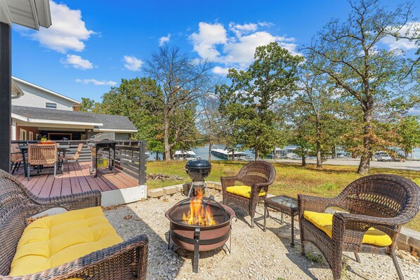 An outdoor patio with wicker furniture, a fire pit, and a view of a lake and buildings in the background. - Table Rock Lakefront Home Near Port of Kimberling_Free Attraction Tickets_Firepit (Kimberling City)