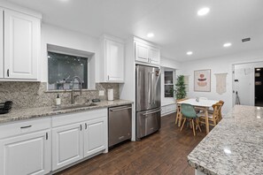 A modern kitchen with white cabinetry, a stainless steel refrigerator, a granite countertop, and a dining area with a table and chairs. - Modern House I Huge Yard I Pool I Cinema & Game Room (Denton)